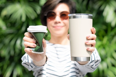 A person holding a crumpled disposable coffee cup and a reusable travel cup towards the camera