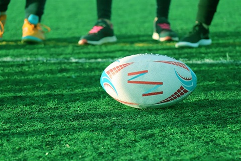 A rugby ball sits on a field with the feet of players standing behind